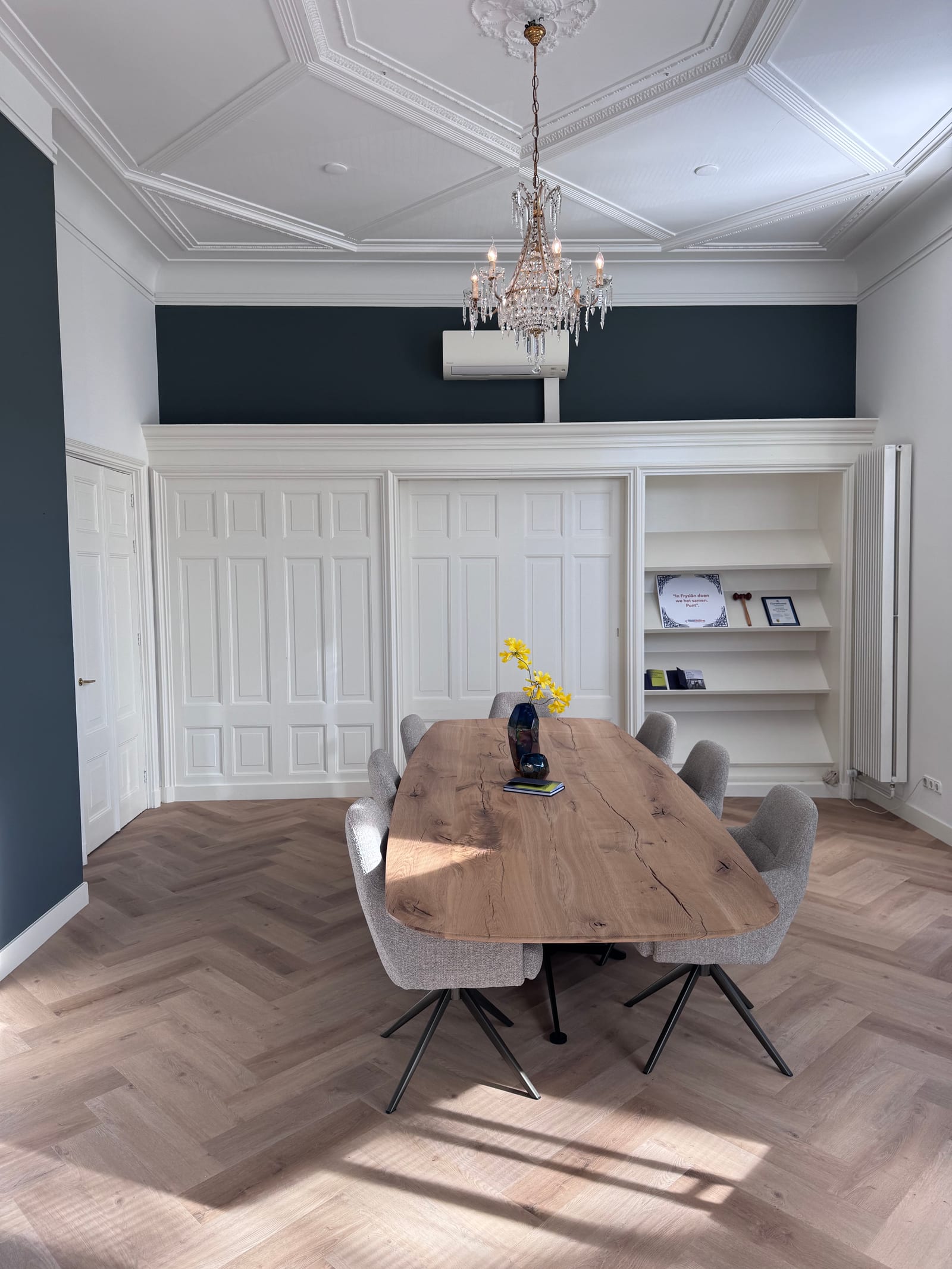 Grote vergaderruimte met een lange eikenhouten tafel, kroonluchter, stucplafond, een witte kastenwand en een donkerblauwe muur.