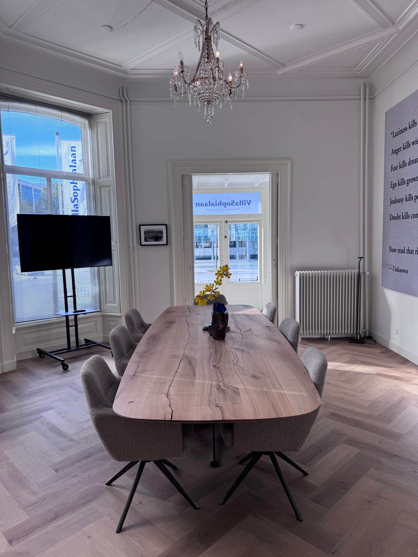 Vergadertafel en grijze stoelen op de visgraatvloer, met de kroonluchter en een doorkijk naar de glazen entree.