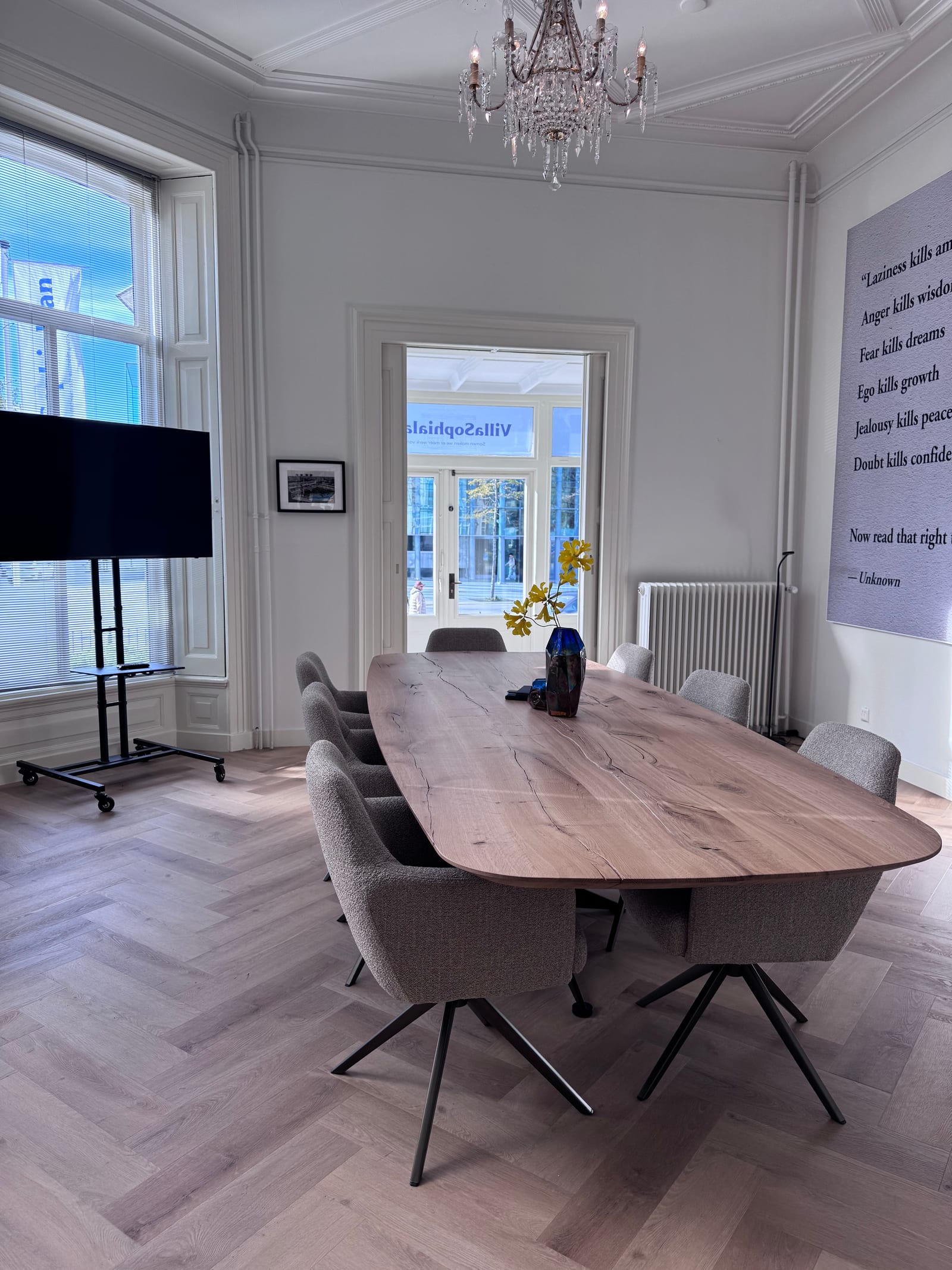 Vergadertafel vanuit de hoek van de ruimte, met hoge ramen, het scherm en de openstaande deur naar de entree.